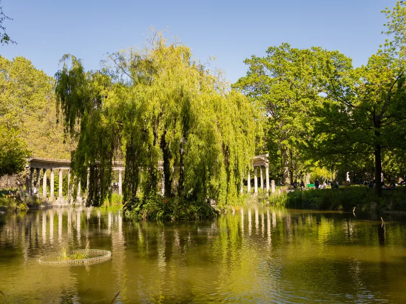 A colonnade and a weeping willow reflected in the pond of a Paris park